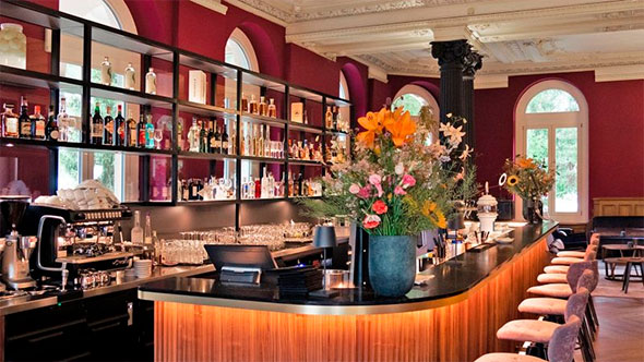 Interior of Vic&rsquo;s Bar featuring a curved bar counter with wooden trim, shelves stocked with alcohol against burgundy walls, and high stools.