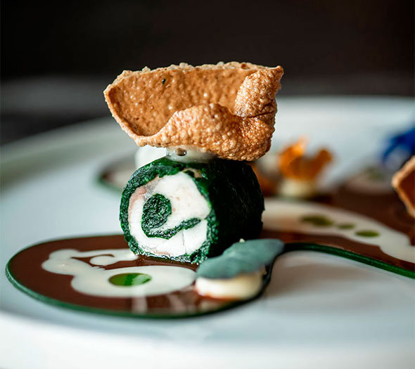 Artistically presented dish with a green roll, a crispy element on top, and sauces on a white plate.