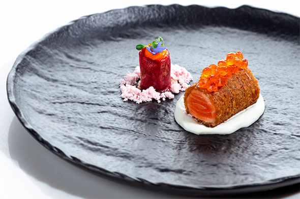 Exquisite salmon roll topped with caviar, served on white sauce and a black textured plate.