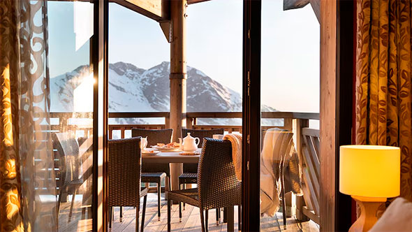 View from inside the apartment onto the balcony with a dining area, opening to a panorama of snow-covered mountain peaks, emphasizing the comfort and ideal location of the Arc 1950 Le Village residence.