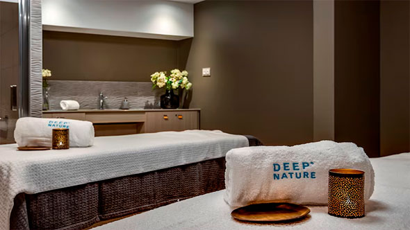 Interior of a Deep Nature Spa treatment room with two massage beds, neatly stacked white towels with the logo, flowers, and candles for relaxing wellness treatments.
