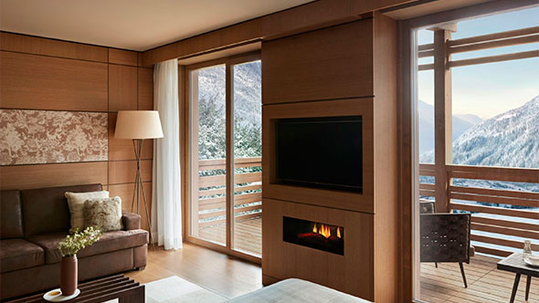 Interior of a luxury hotel suite (Prestige Junior Suite, Lefay) with wood paneling, fireplace, and panoramic balcony view of the snowy Dolomites.