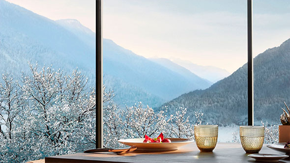 An elegant dining setting with a gourmet dish and golden glasses on a wooden table, featuring a panoramic window view of the snow-covered Dolomite mountains, highlighting the hotel’s gastronomic experience.