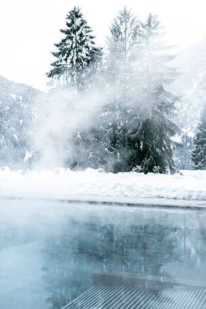 A view of the outdoor heated Lefay SPA pool with steam rising, set against a snow-covered Dolomite landscape and tall pine trees, capturing the essence of winter wellness.