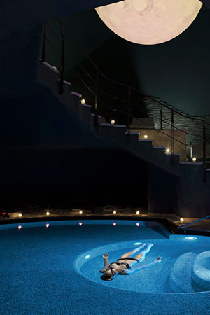 A woman floating on her back in an illuminated pool under a large moon image, creating a serene night ambiance in a luxurious Lefay Resort SPA with a candle-lit staircase.
