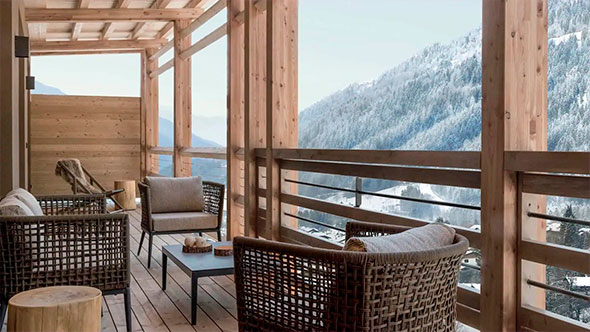 Large wooden balcony of a luxury chalet (Lefay style) with rattan outdoor furniture, offering a panoramic view of a snowy mountainside and ski slope.