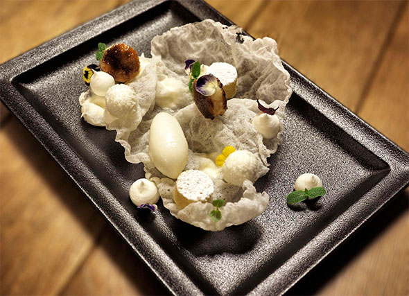 A refined dessert on a black plate, featuring white sorbet or mousse, a crispy rice chip, mini meringues, coconut flakes, caramelized elements, and violet flowers for decoration.