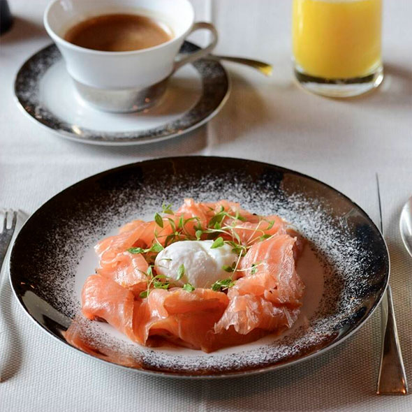 Exquisite breakfast or brunch: thinly sliced smoked salmon arranged in a circle around a poached egg, garnished with microgreens, served on a black plate with a white rim, with a cup of coffee and a glass of orange juice nearby.