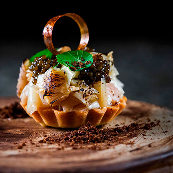 Exquisite starter served in a tartlet: filling made of caramelized leeks or vegetables topped with black caviar, garnished with a leaf and a caramel ring, presented on a rustic wooden or ceramic plate dusted with crumbs.