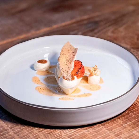 An exquisite dessert on a wide white plate with a grey rim: a central element of vanilla ice cream or mousse with caramelized grapefruit and a thin wafer chip, surrounded by sauce and two small portions of cream.