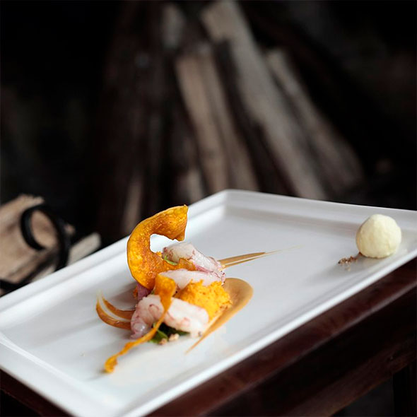 A fine dining dish on a white rectangular plate: langoustines or shrimp served with a bright pumpkin purée, thin spirals of roasted pumpkin, and a savory sauce, with a sphere of creamy mousse or sorbet on the side.