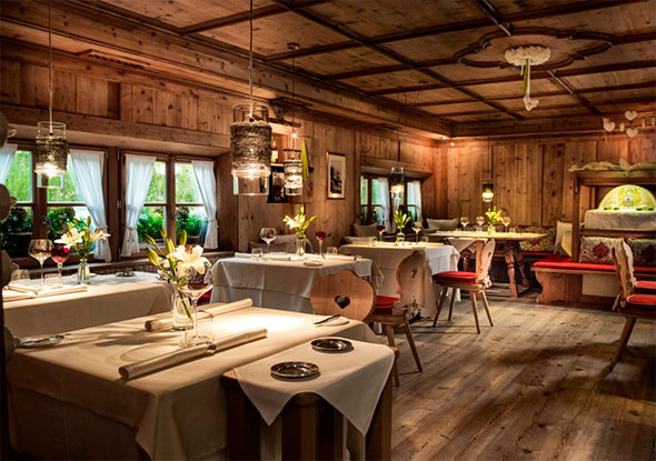 Cozy Michelin Stube restaurant interior in Alpine style: walls and ceiling paneled with natural wood, white tablecloths, chairs with red folk-style cushions, and soft lighting perfect for a romantic dinner.
