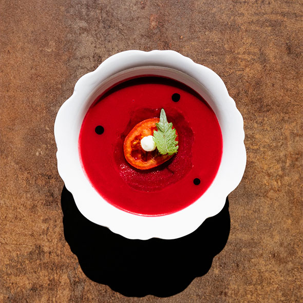 Vibrant gazpacho or beetroot purée soup (vegetarian dish) in a white scalloped bowl, garnished with a slice of charred tomato, a sphere of cream (sour cream/mousse), and a fresh mint leaf, set against a dark wooden background.