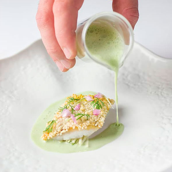 The process of serving a refined dish: a chef’s hand pours a delicate green sauce from a pitcher over a fillet of crispy-crusted fish (possibly with herbs or seeds) onto a textured white plate.