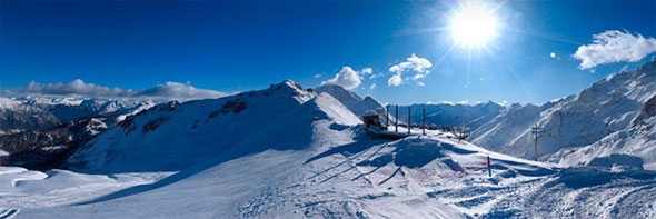 Panoramic view of the snow-covered high-altitude slopes of Serre Chevalier under bright sun, showing wide ski runs, a lift station, and the majestic Écrins Massif in the background.