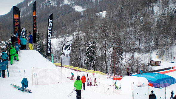 A lively snowpark in Serre Chevalier with skiers and snowboarders practicing tricks on jumps and landing pads, surrounded by snow-covered forest and branded flags