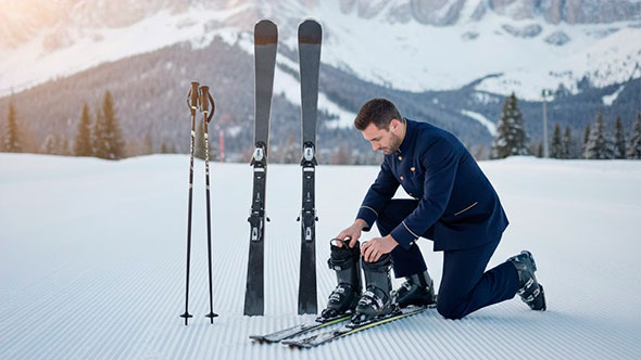 Ski butler or assistant in uniform kneeling on a pristine snowy slope, preparing ski equipment (skis and boots) with Dolomites mountains in the background.