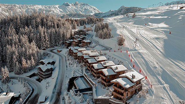 Aerial view of a row of luxury Ultima Collection chalets in Megève, situated along a road and near the ski slopes, overlooking the snow-covered Alps and majestic peaks.