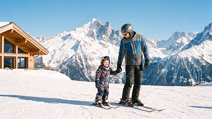 Parent and small child in helmets on the snow below the peaks of the French Alps